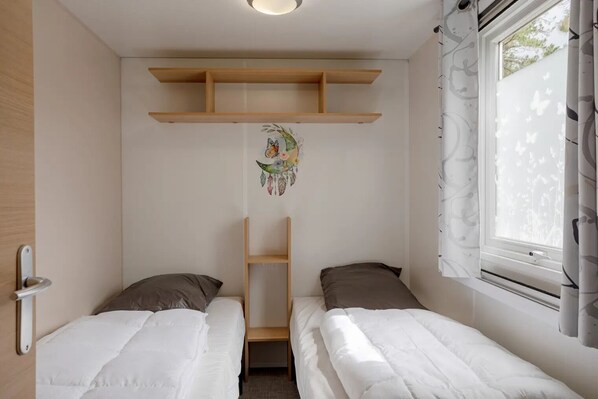 Ein kleines, sauberes Zimmer mit zwei Einzelbetten, einem Holzregal darüber und einem Fenster mit Vorhängen.