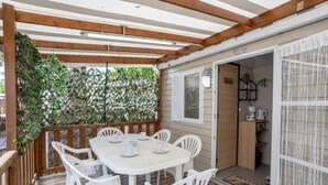 Eine überdachte Terrasse mit Tisch und Stühlen, ein Holzdeck, eine Tür zu einer Küchenzeile und Blick ins Grüne durch einen Spalierzaun.