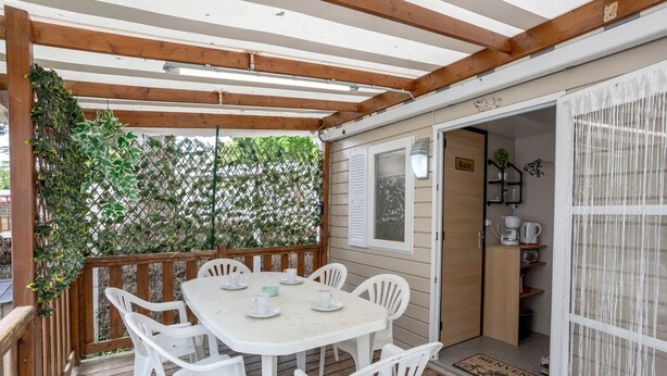 A covered patio with a table and chairs, a wooden deck, a door leading to a kitchen area, and a view of greenery through a lattice fence.