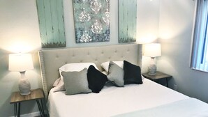 Un lit bien fait, recouvert d’une couette blanche et agrémenté de trois oreillers dans des tons de gris et de noir. On y trouve deux tables de chevet avec des lampes, et un élément décoratif mural au-dessus du lit.