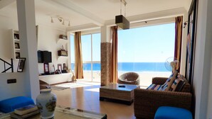 Una sala de estar con vista a la playa, muebles de mimbre, una mesa de centro de vidrio y un televisor.
