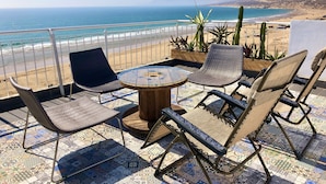 Una terraza con una mesa redonda y seis sillas, con vistas a una playa que tiene una valla y plantas.