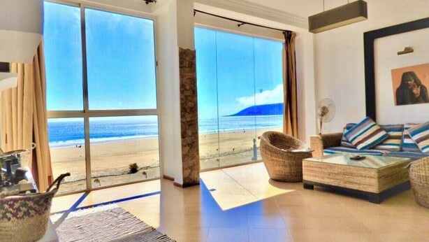 Un salón espacioso con un amplio ventanal corredera que da a la playa, muebles de mimbre y un cuadro en la pared.