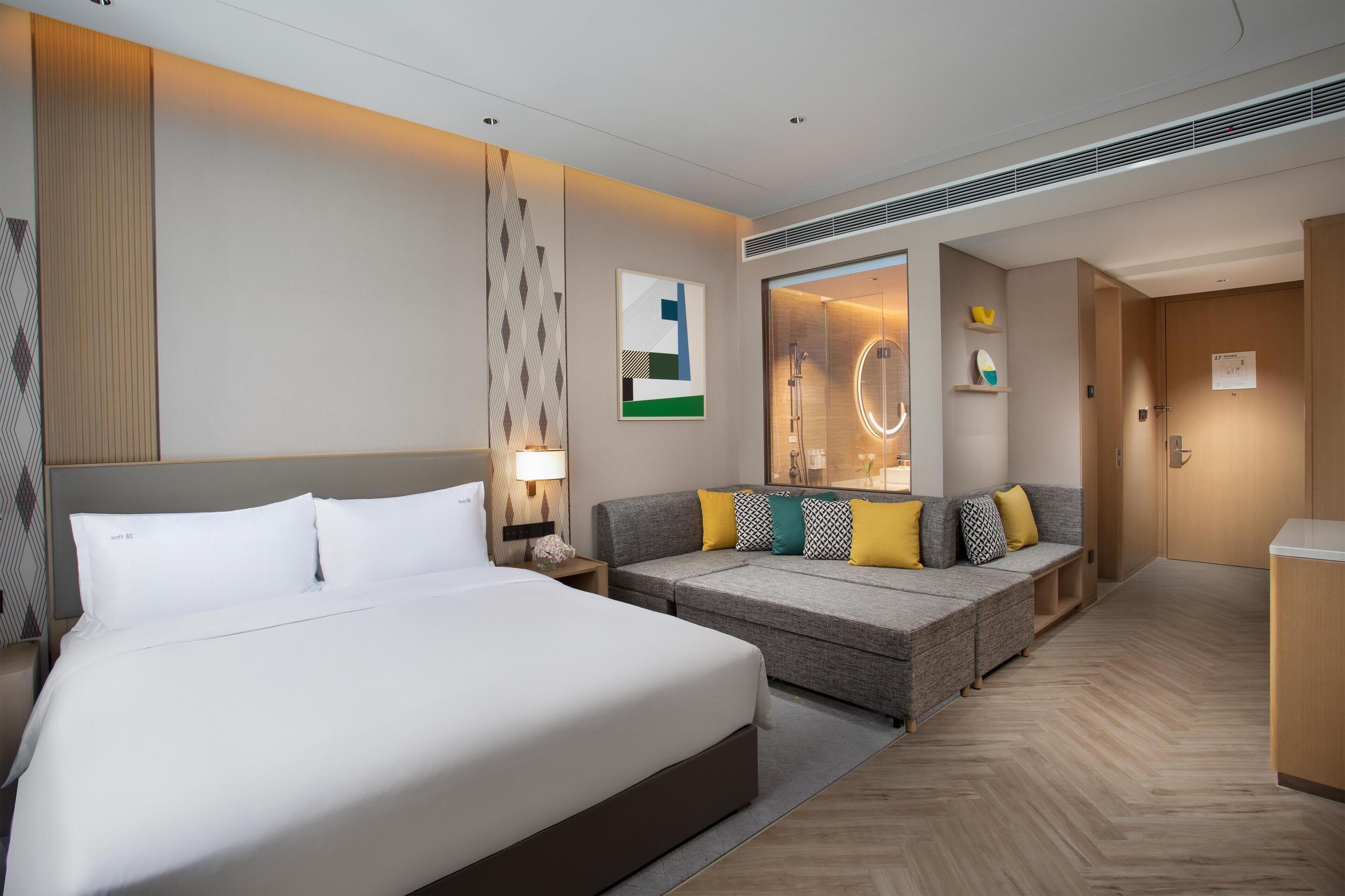 A modern hotel room with a large bed, a sofa, a mirror, and a small kitchenette.