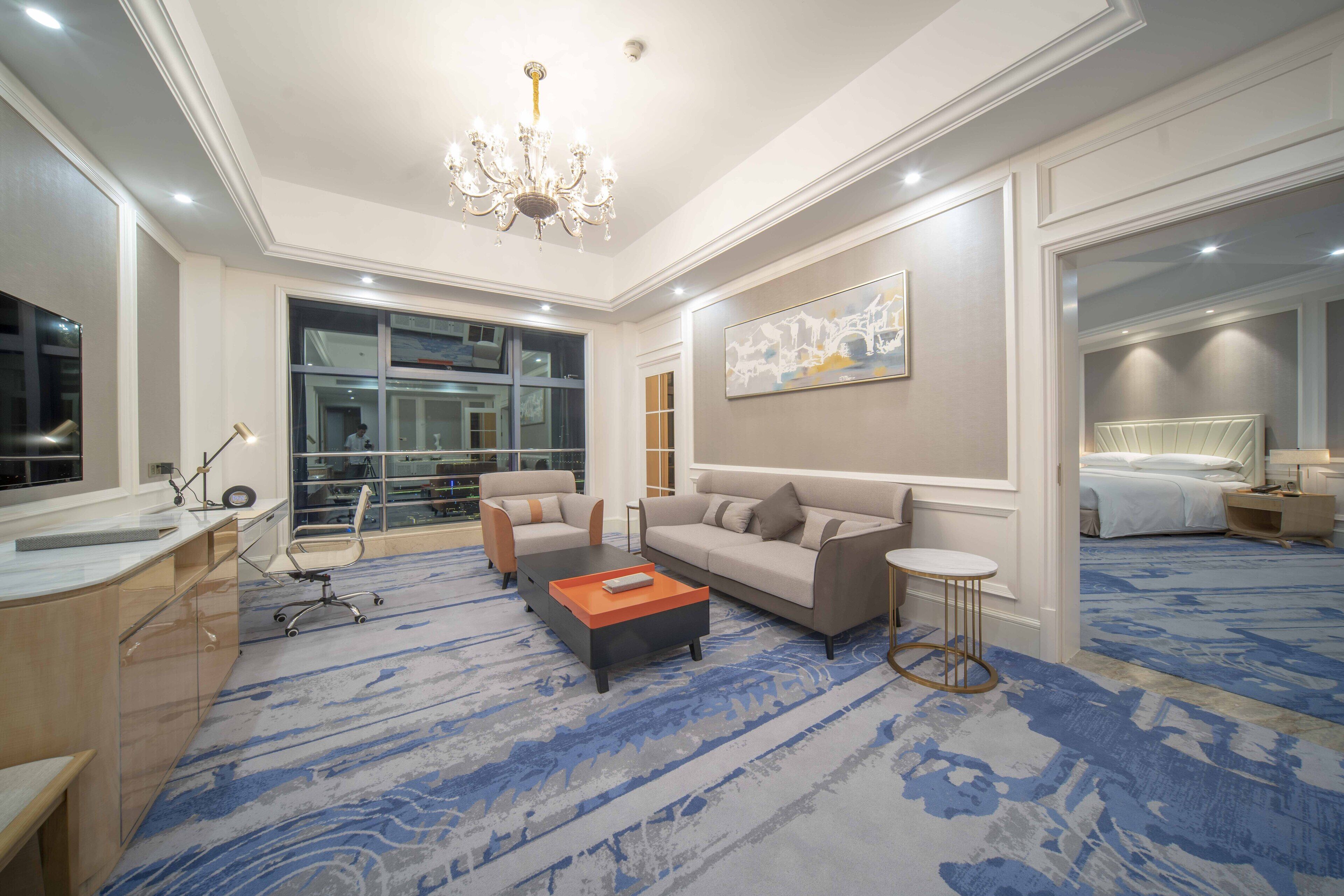 A hotel room with a large window, a chandelier, a sofa, a coffee table, and a bed.