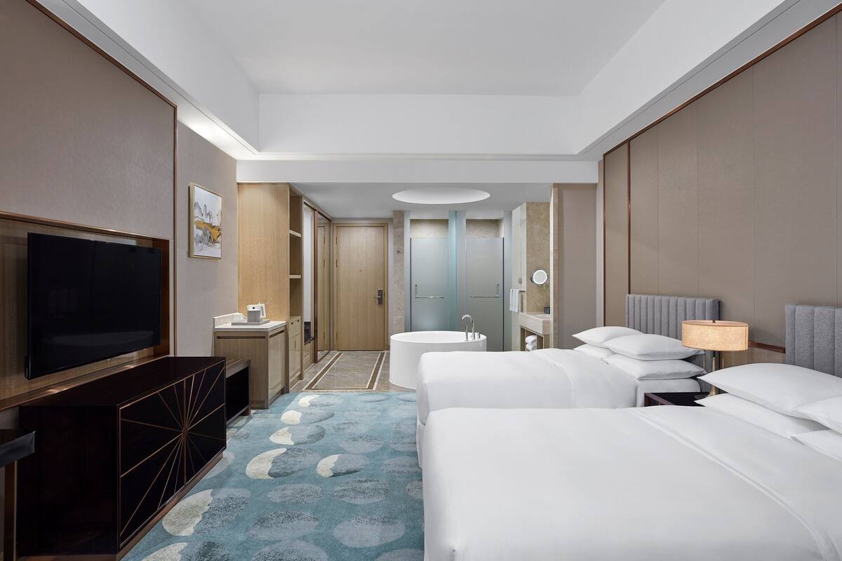 A hotel room with a TV, two beds, a desk, a bathroom, and a large mirror.