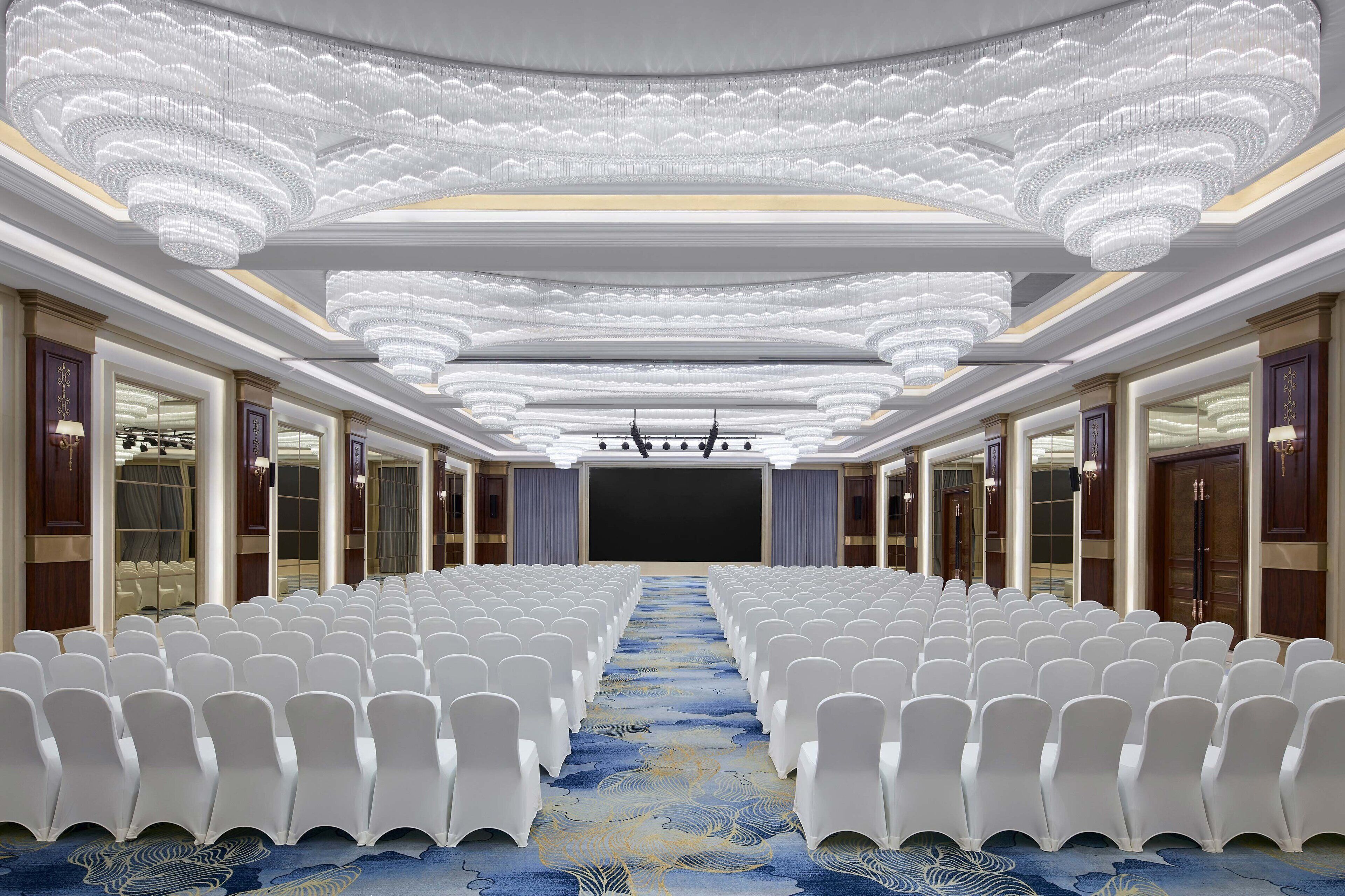 A conference room with rows of white chairs, a blue carpet, and ornate chandeliers.