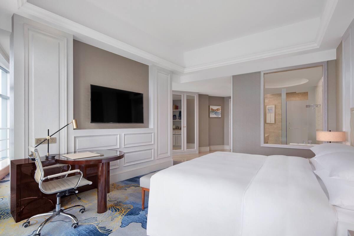 A hotel room with a large bed, a desk with a chair, a flat-screen TV mounted on the wall, and a bathroom visible through an open door.