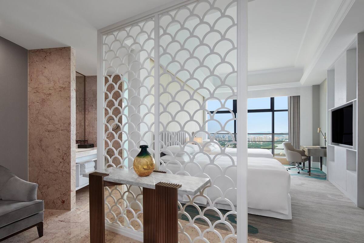 A modern hotel room with a large bed, a desk with a chair, a TV, and a view of the city.