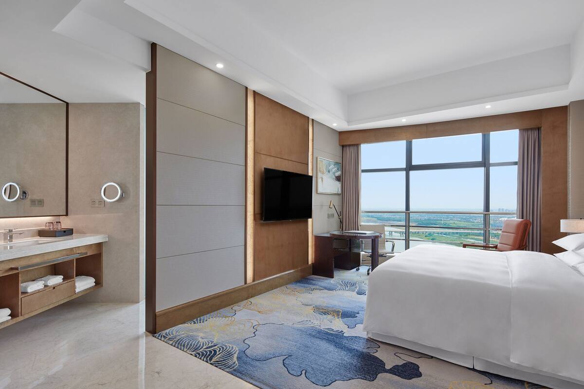 A modern hotel room with a large bed, a bathroom with a vanity, a flat-screen TV, and a view of the city.