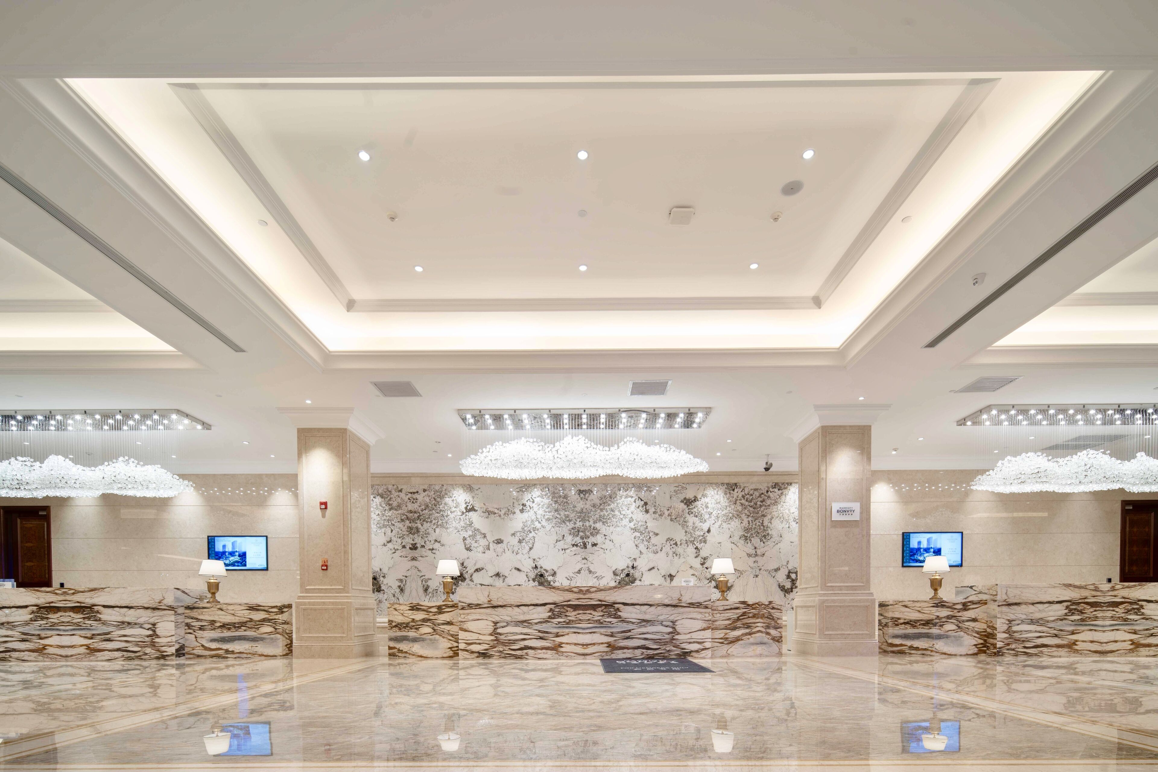 A spacious lobby with a marble reception desk, modern chandeliers, and a high ceiling with recessed lighting.