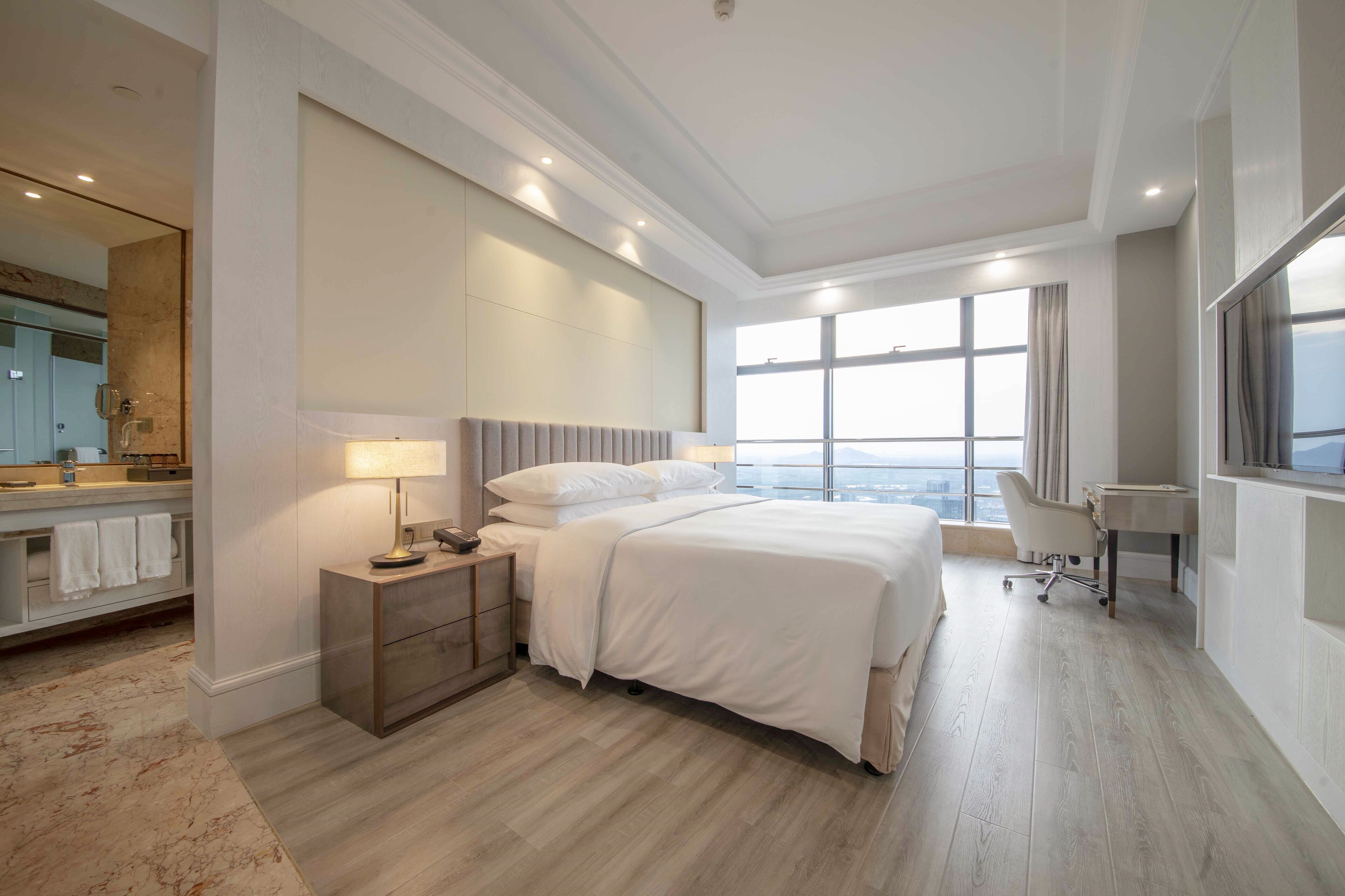 A modern hotel room with a large bed, a desk, a chair, and a view of the city.