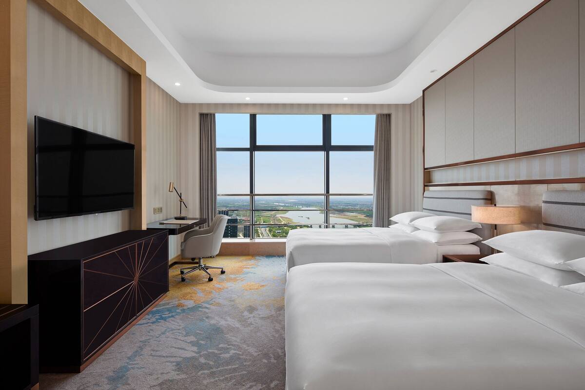 A hotel room with two beds, a desk, a TV, and a large window offering a city view.