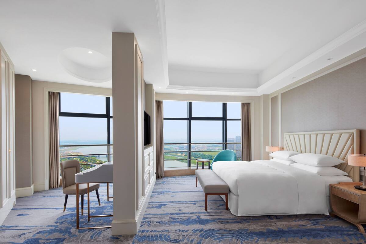 A hotel room with a large bed, a desk, a chair, and a view of a cityscape through large windows.