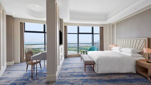 Habitación de hotel con una cama grande, un escritorio, una silla y vistas a un paisaje urbano a través de amplios ventanales.