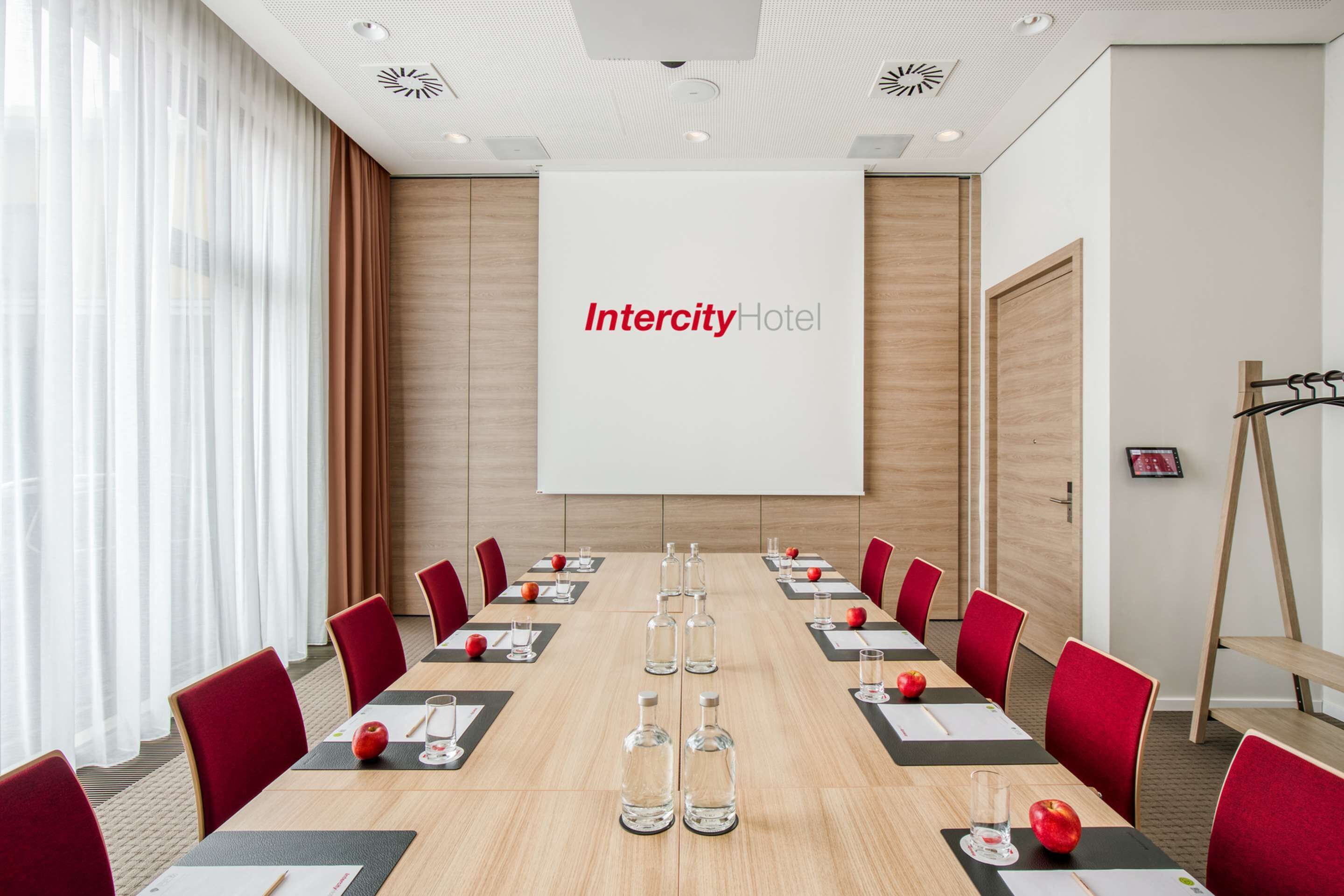 Un salón de conferencias con una larga mesa, sillas rojas y una pantalla de proyector que muestra 'Intercity Hotel'.