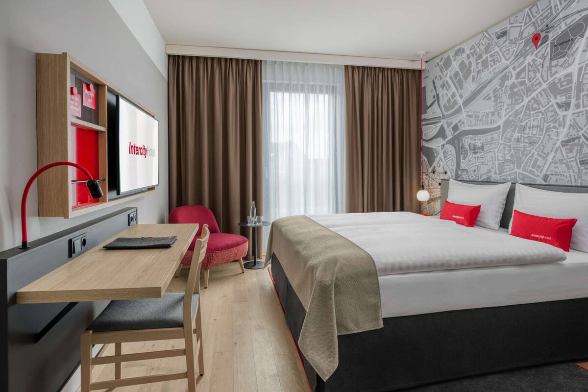 A hotel room with a bed, a desk, a chair, a small table, and a map on the wall.