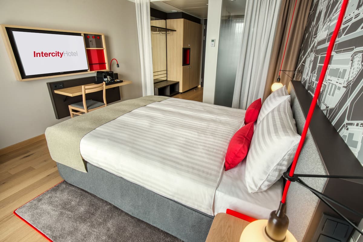 A modern hotel room with a bed, a desk, a TV screen displaying "Intercity Hotel," and a cityscape wall art.