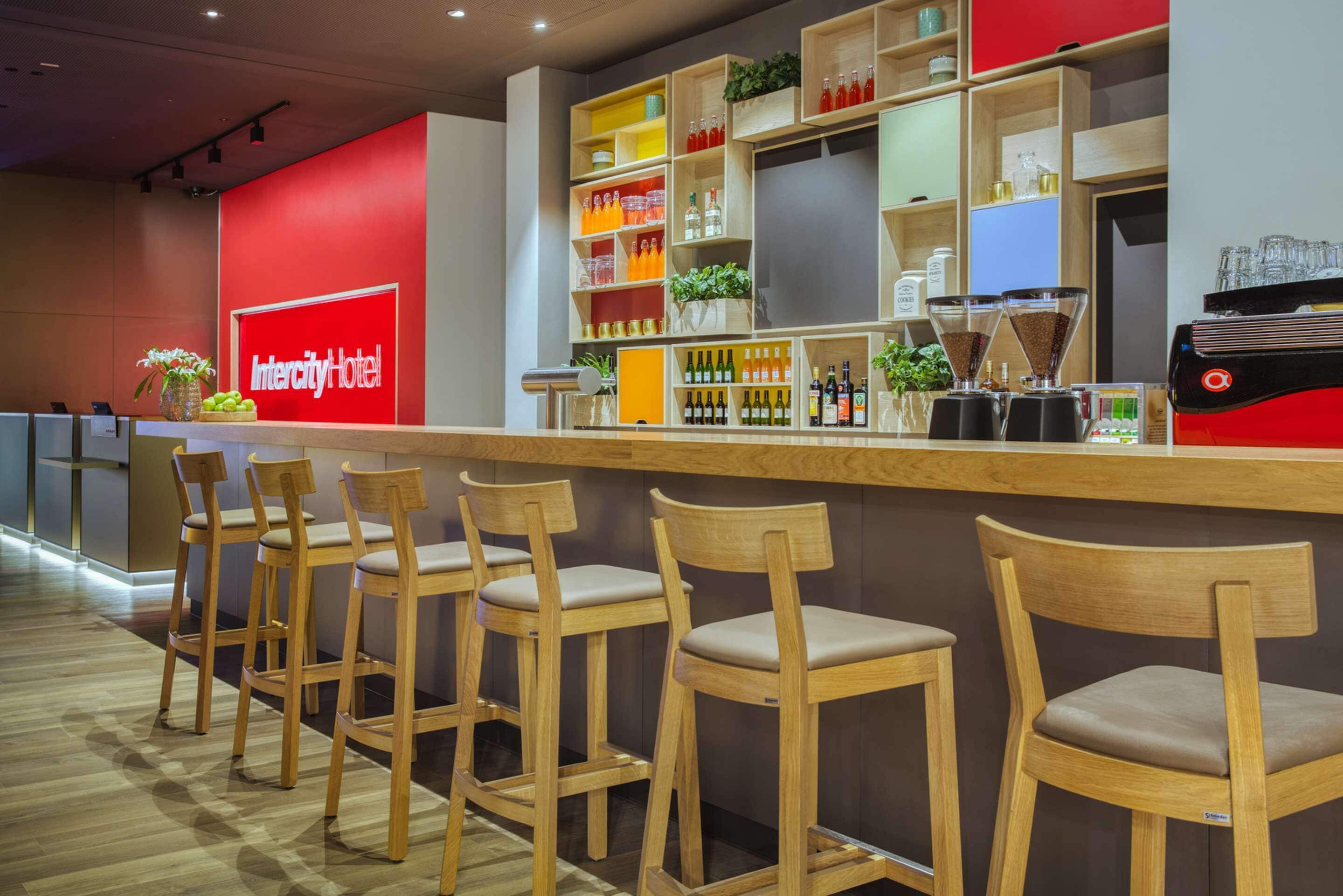 Un bar d’hôtel moderne, avec des tabourets de bar en bois, un mur rouge orné du nom de l’hôtel, et des étagères remplies de bouteilles et d’ustensiles pour préparer le café.