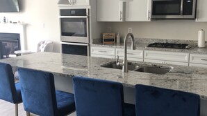 Cocina moderna con armarios blancos, horno empotrado, microondas, pileta y encimera de mármol. Cuatro taburetes azules y una lámpara colgante.