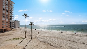 A beach with a building on the left, palm trees, and the ocean. - Ocean Views From Every Window | Condo 206C at Estero Beach & Tennis Club + 2 Bikes, Beach Gear. 6-7night stays (Fort Myers Beach)