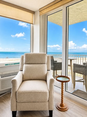 A room with a large window overlooking a beach, a beige armchair, wicker chairs, and a small round table.