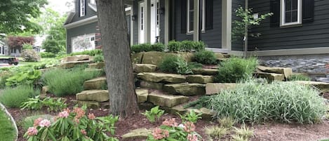 A house with a front porch, a tree, and a landscaped garden with shrubs and flowers.