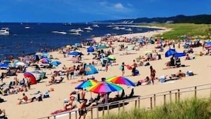 Een druk strand met mensen die zonnebaden en zwemmen, en boten op het water.