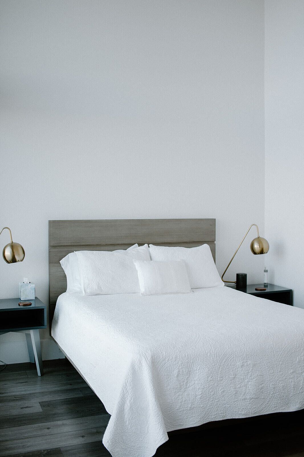 A neatly made bed with a white bedspread and pillows, flanked by two bedside tables with lamps, in a minimalist room with a wooden floor.