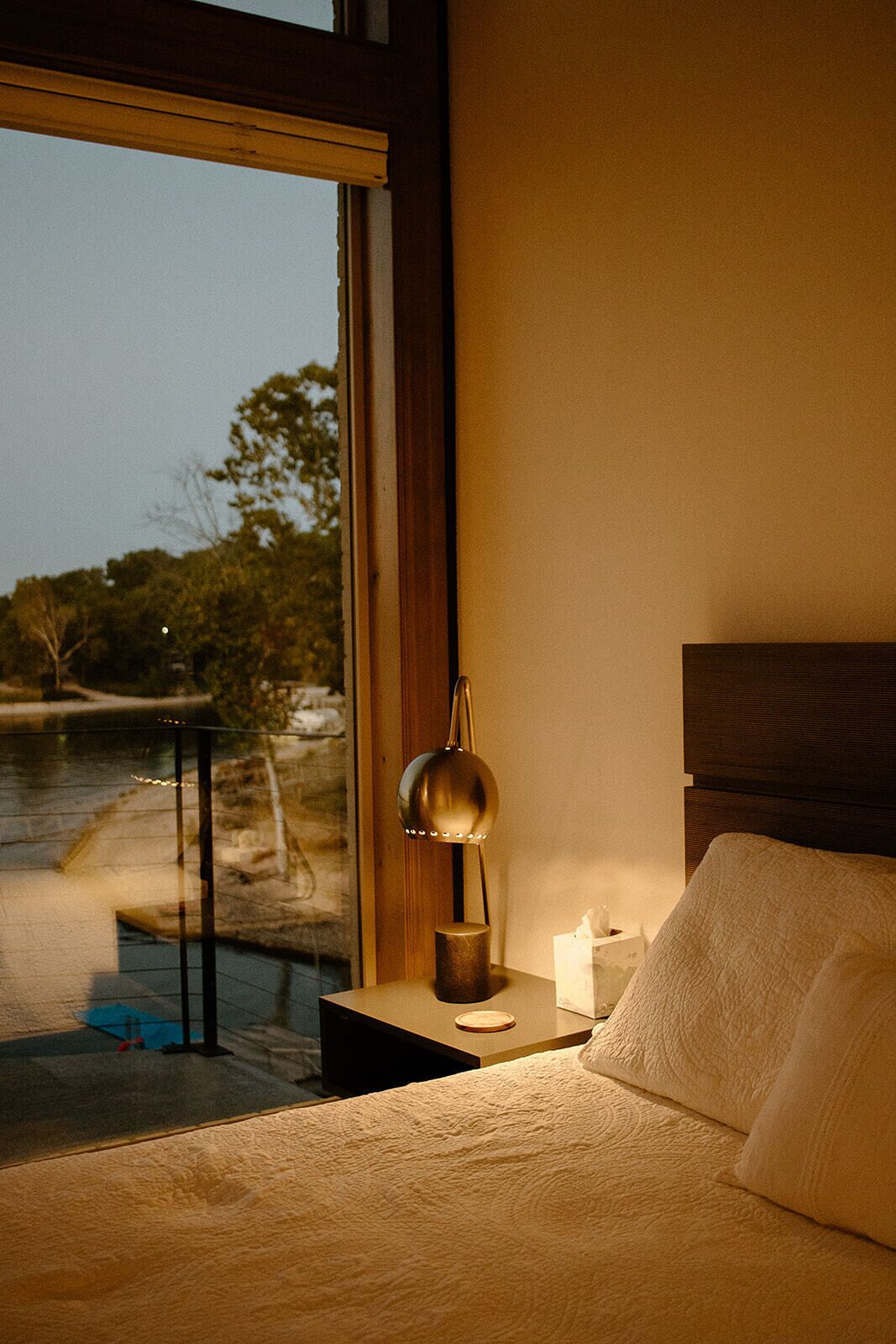A bedroom with a large window, a bedside table with a lamp, a bedside table with a tissue box, and a bed with pillows.