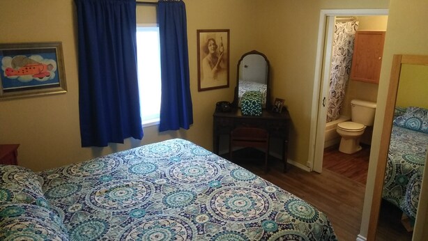 A bedroom with a bed, a dresser, a mirror, a window with curtains, a painting, and a bathroom visible through an open door.
