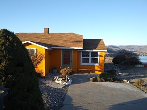 A yellow house with a brown roof, a small front porch, and a view of a lake and mountains in the background.