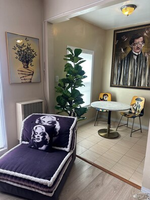 A room with a bed, a potted plant, a dining table with chairs, and a large painting on the wall.