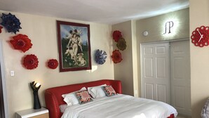 A bedroom with a red bed, white bedding, a nightstand, a red chair, a door, and decorative glass flowers on the wall. - Milano Art Studio Apartment (Los Angeles)