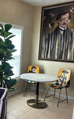 A dining area with a round table, two chairs, a potted plant, and a large painting on the wall.