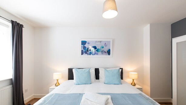 A hotel room with a bed, two bedside lamps, a window with curtains, a wall-mounted artwork, and a towel on the bed. - Velvet 1-bedroom Apartment The Clockhouse, Hoddesdon (Hoddesdon)