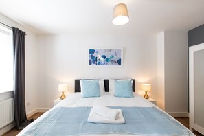 Una habitació d'hotel amb un llit, dues làmpades de nit, una finestra amb cortines, una obra d'art penjada a la paret i una tovallola sobre el llit.
