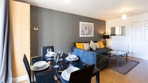 A modern living room with a dining area, grey walls, a sofa with cushions, a glass coffee table, and a framed picture on the wall. - Velvet 1-bedroom Apartment The Clockhouse, Hoddesdon (Hoddesdon)
