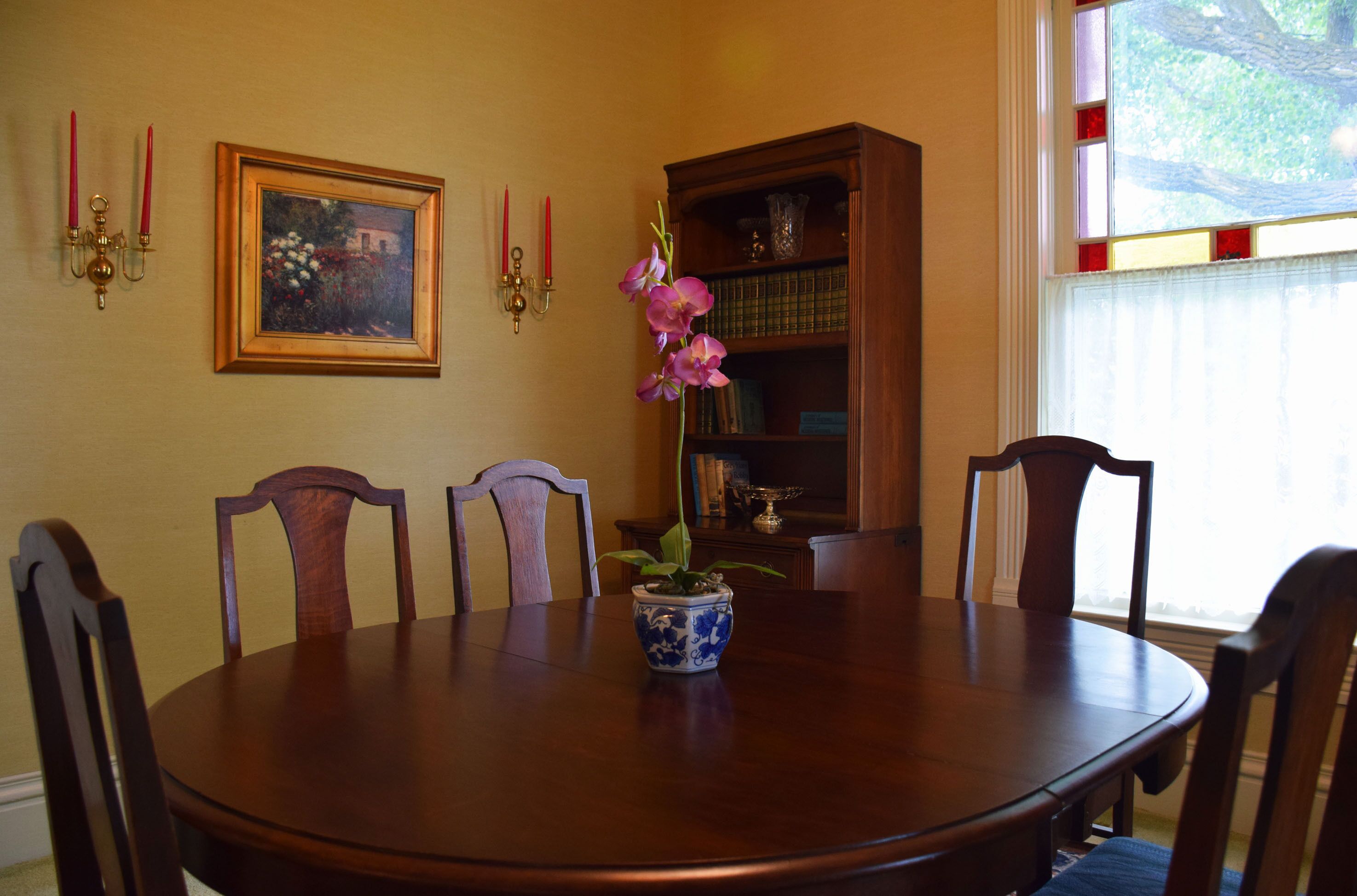 A dining room with a round wooden table, six wooden chairs, a vase with flowers, a framed painting, a bookshelf, and two wall sconces with candles.