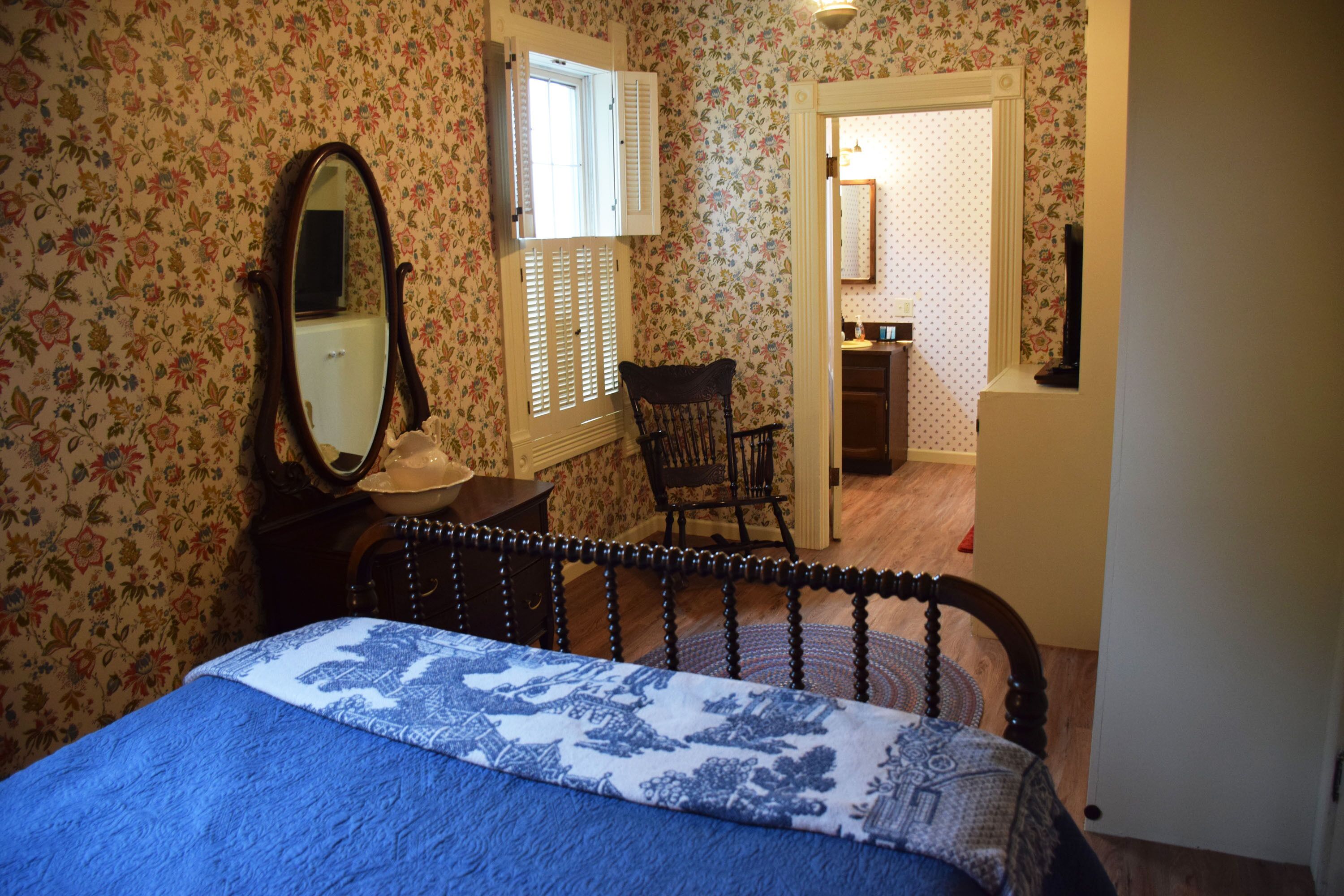 Un dormitorio con papel tapiz floral, una cama con colcha azul y blanca, una silla de madera, un aparador y un espejo.