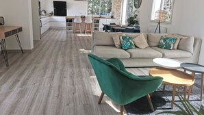 Una sala moderna con sofá, sillón verde y mesa de centro, que da paso a una cocina y zona de comedor con luz natural y una pared de piedra decorativa.