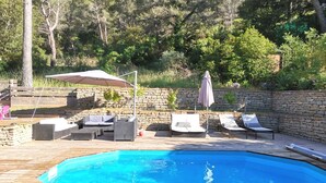 Una piscina con sillas de descanso y sombrillas, rodeada por un muro de piedra y vegetación.