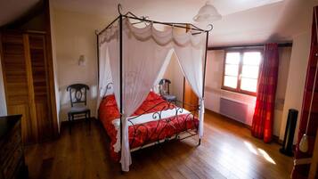 Un lit à baldaquin à quatre poteaux, avec une couvre-lit rouge et un ciel de lit blanc, un plancher en bois, une chaise, une petite table et une fenêtre avec des rideaux.