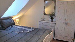 Une chambre avec un plafond en pente, un lit recouvert d’une couvre-lit à rayures, une table de chevet avec une lampe, une commode, une armoire et un petit tapis.
