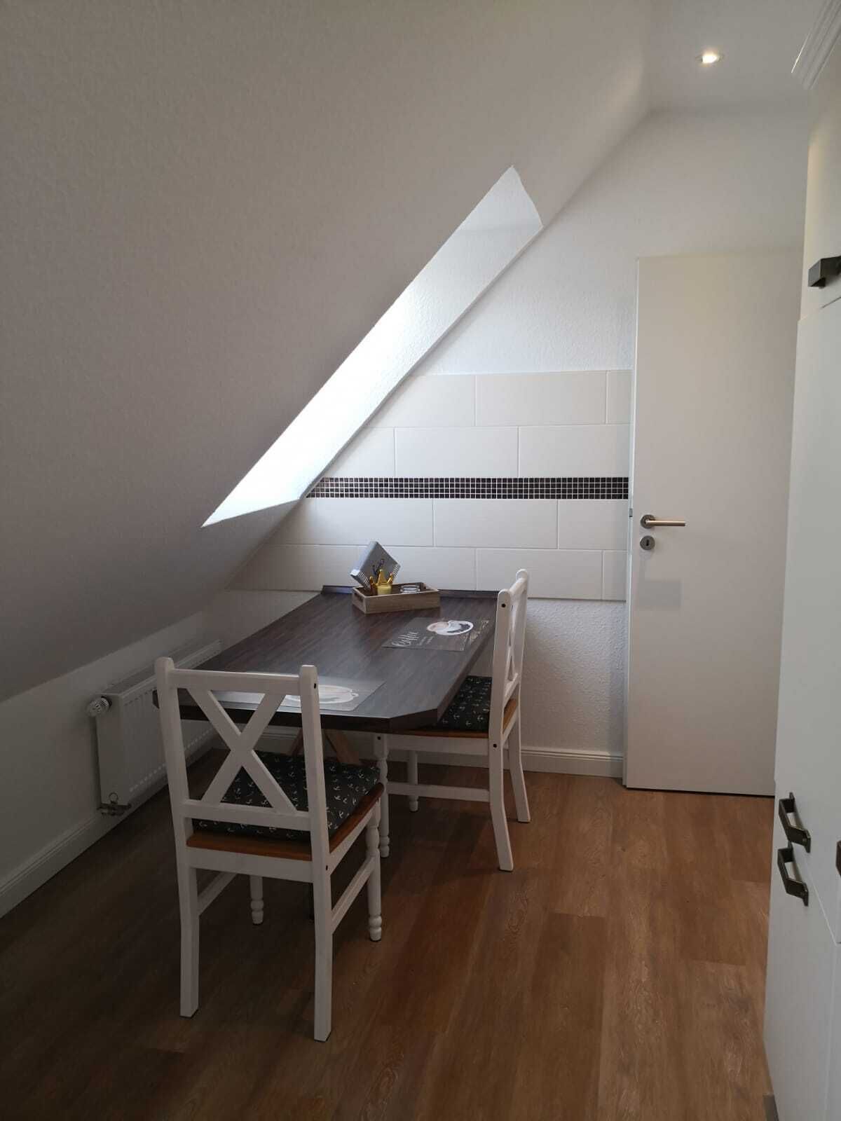 Un petit coin repas avec une table en bois foncé et des chaises blanches, un mur recouvert de carreaux et un plafond en pente.