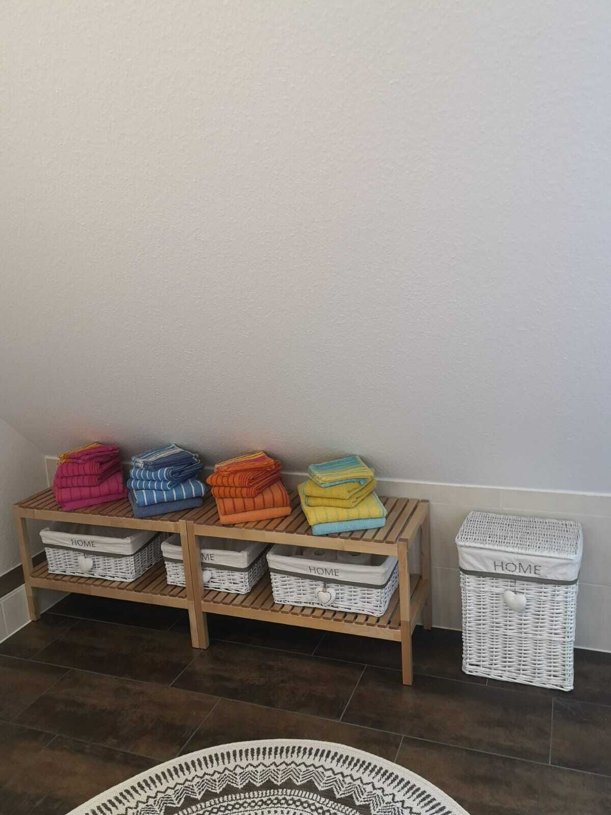 Un angolo con uno scaffale di legno che contiene coperte piegate in vari colori, due cesti in vimini con l'etichetta 'HOME' e un cesto in vimini bianco con un oggetto bianco sopra.