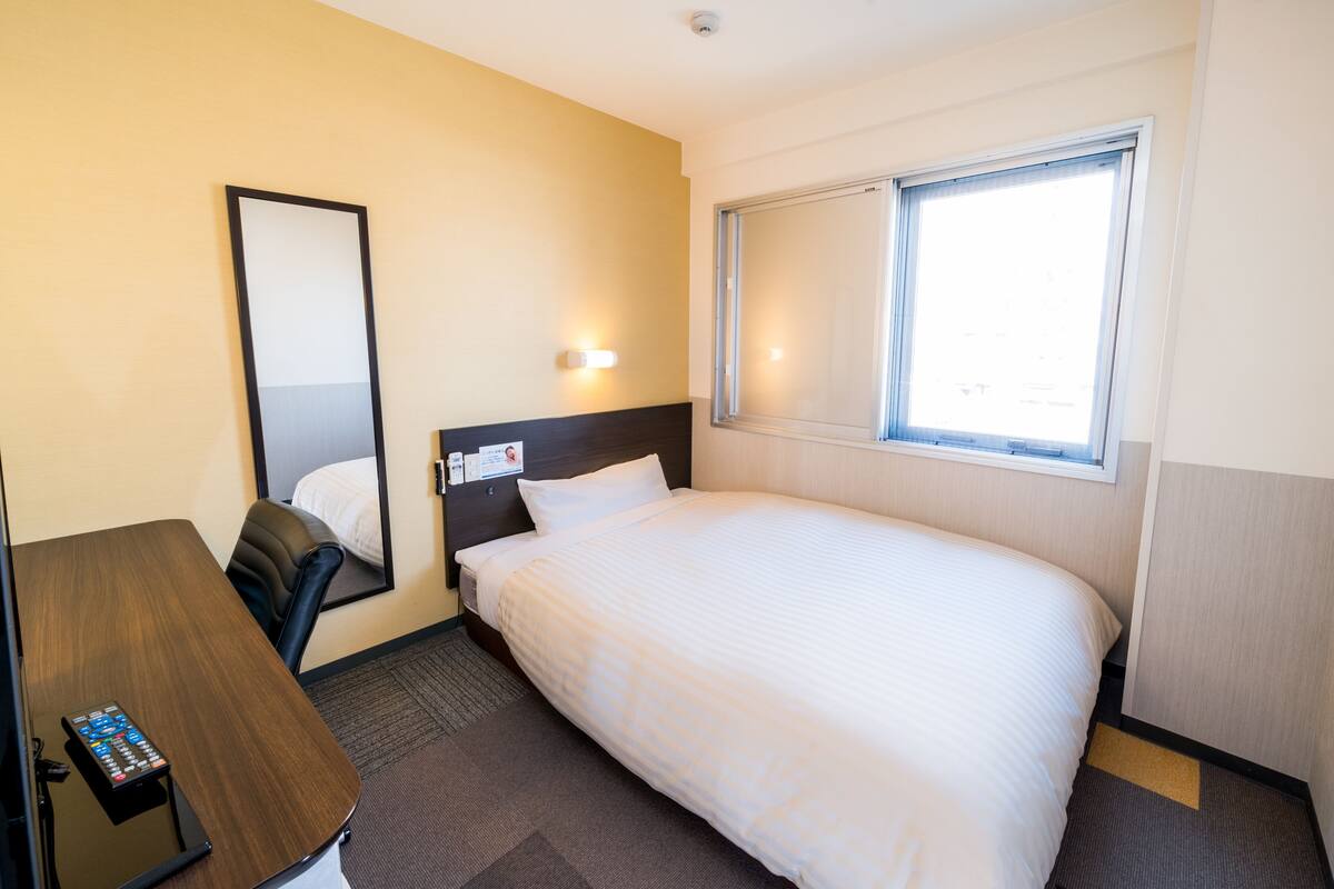 A hotel room with a bed, a desk, a chair, a mirror, and a window.