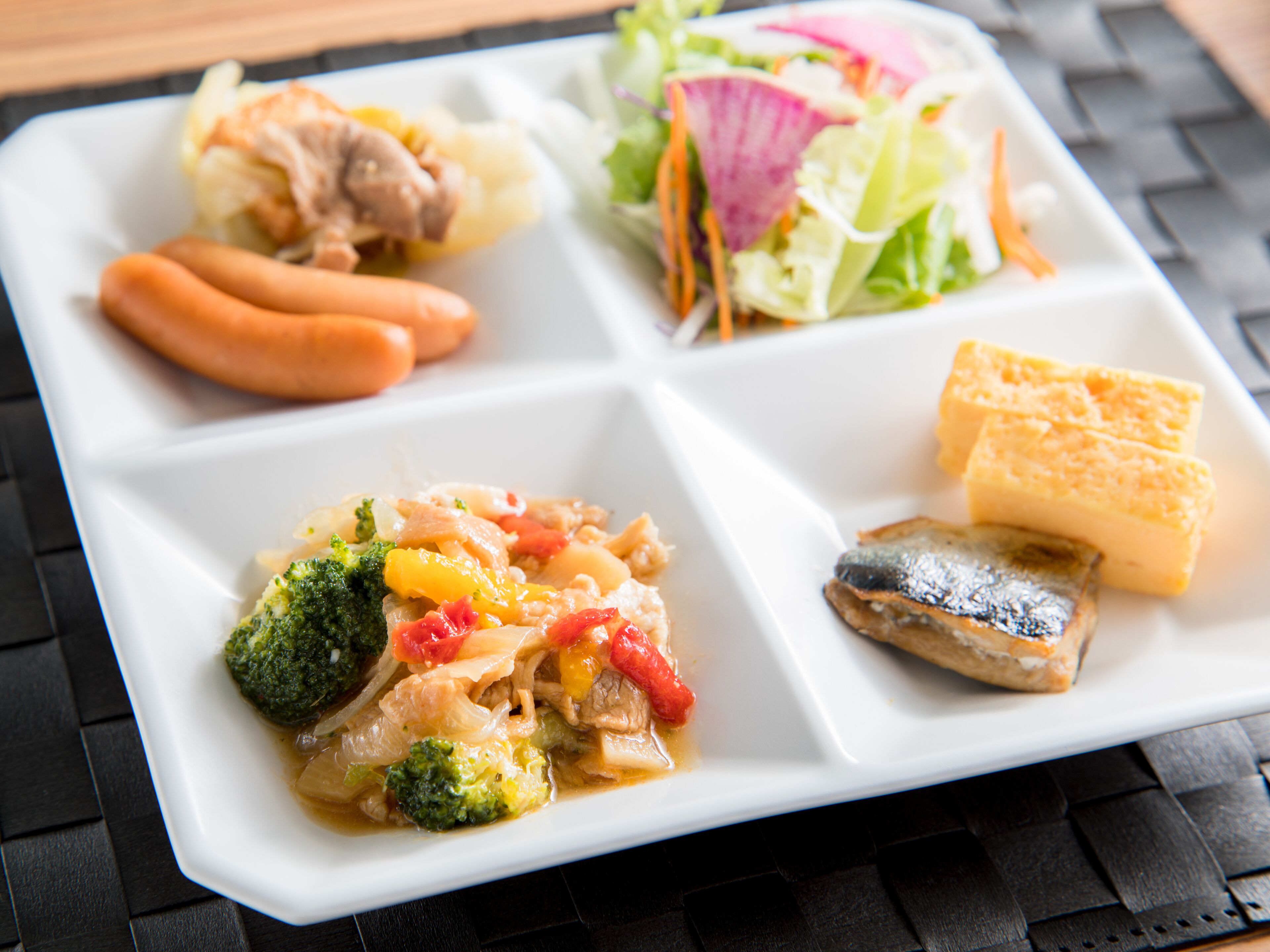 A white rectangular plate divided into four sections with various foods, including sausages, a salad, tofu, and a fish.