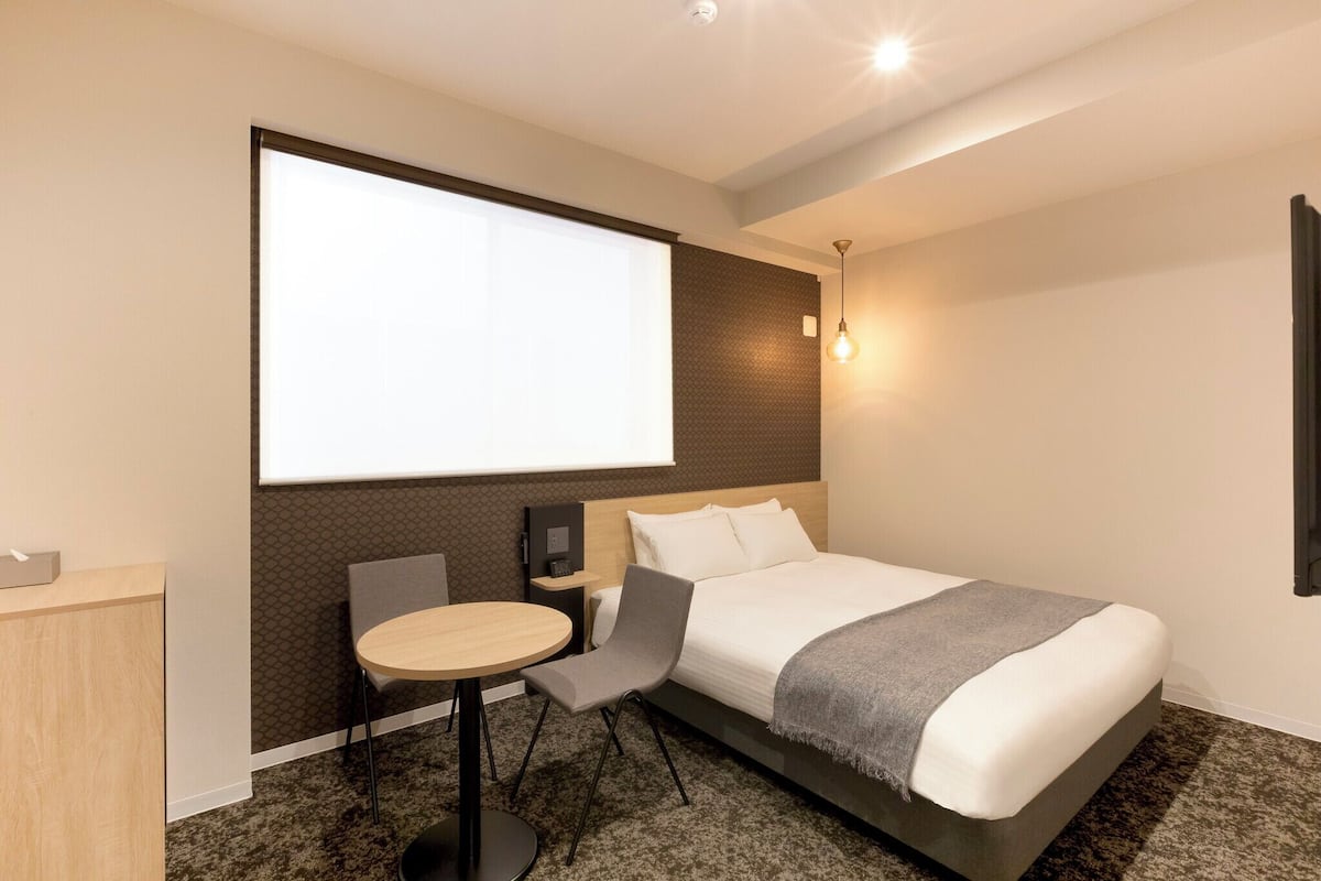 Quarto de hotel moderno com uma cama grande, uma mesa, uma cadeira e uma tela de projeção.