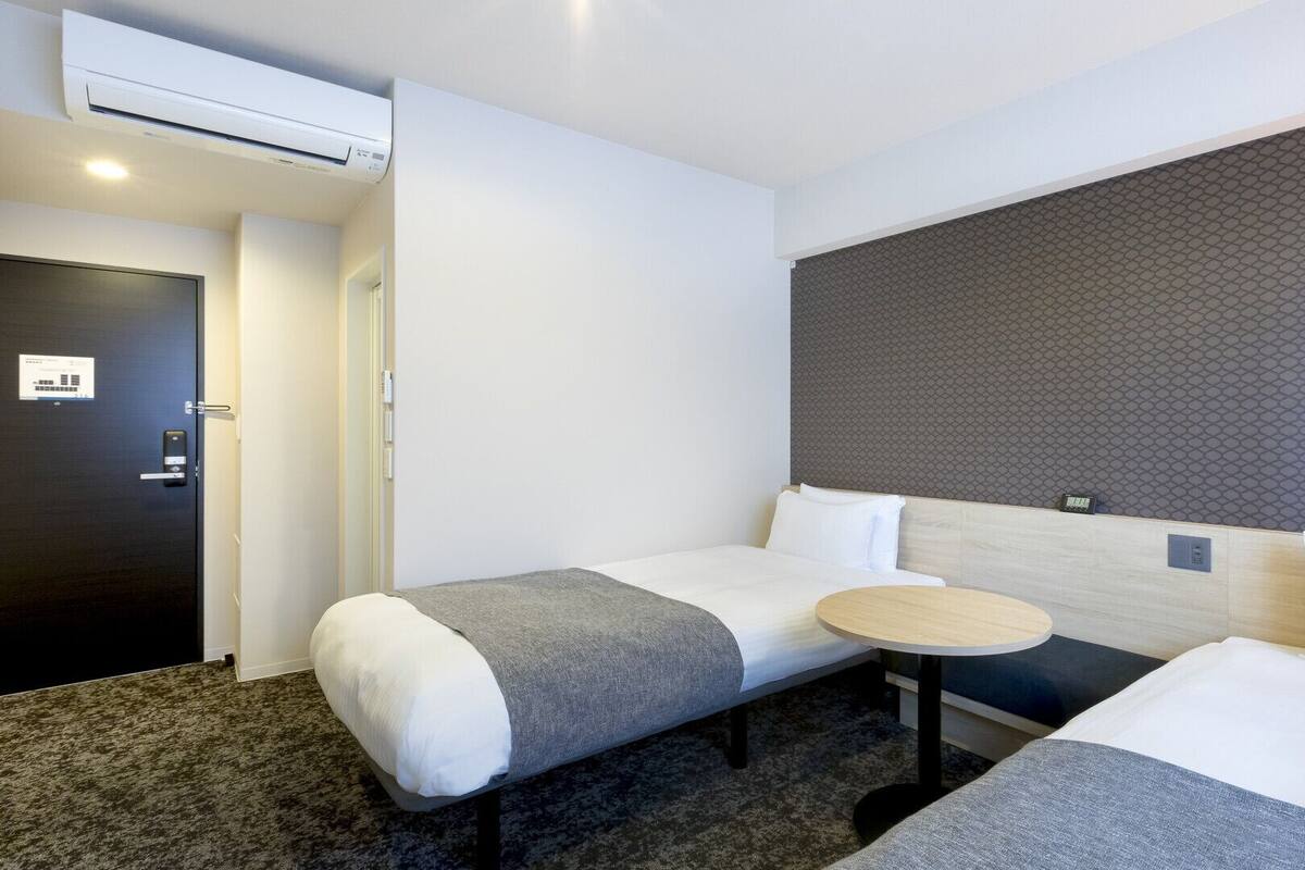 Quarto de hotel com duas camas, uma mesa redonda pequena, um ar condicionado e uma porta com tranca.
